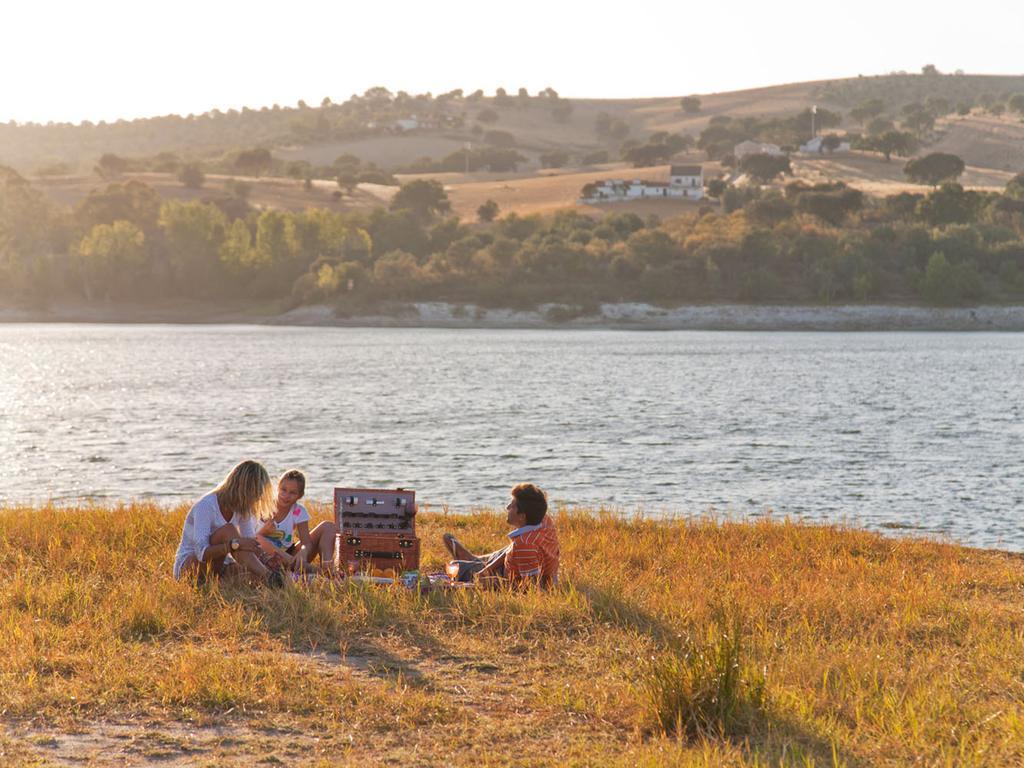 Ap Lago Montargil - Conference & Spa Exterior photo The photo depicts a serene lakeside scene during sunset. Three people are gathered on a grassy area near the water, enjoying a picnic. They are seated around a picnic basket, with the shimmering lake in the background and hills in the distance. The w