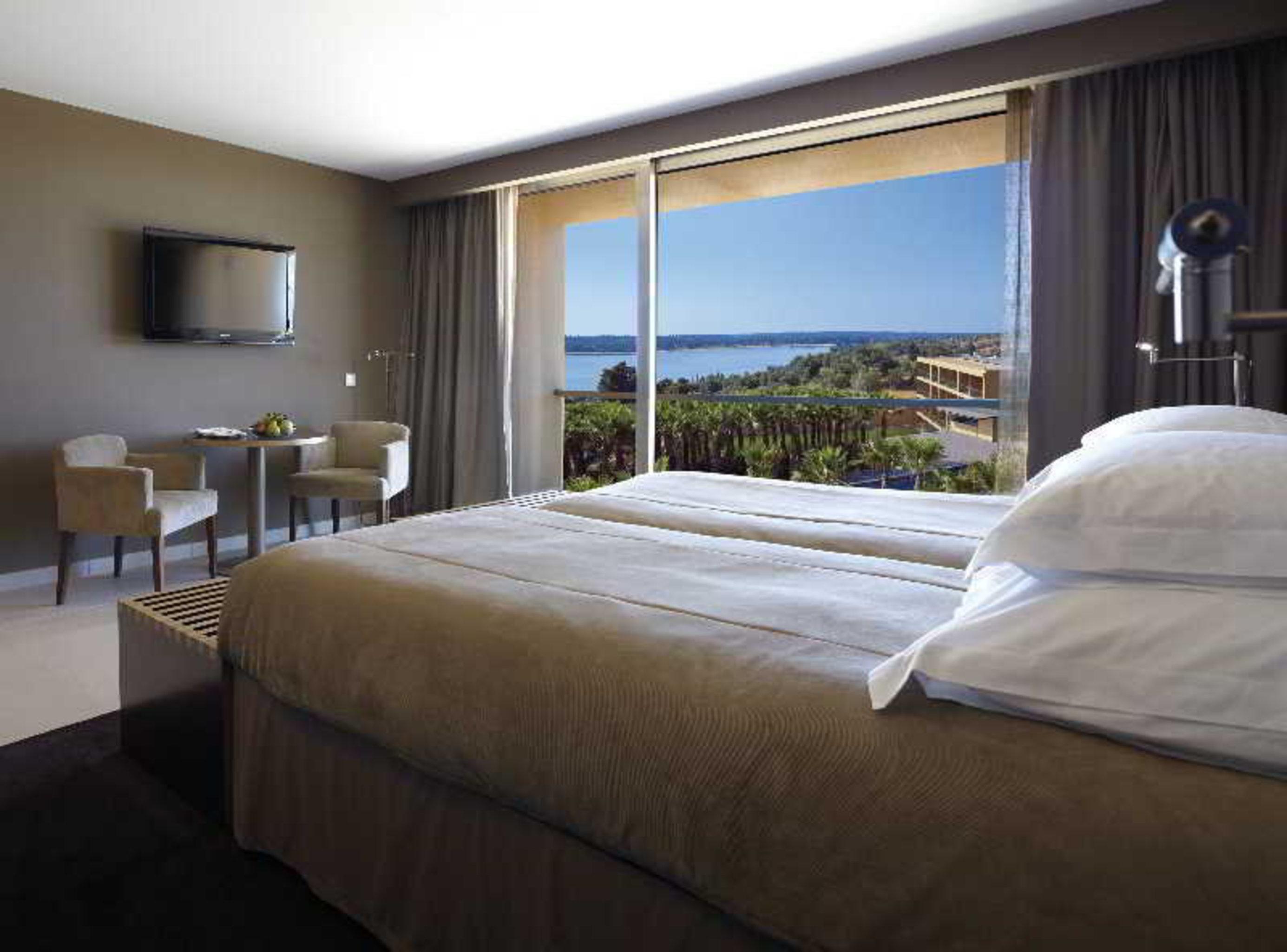 Ap Lago Montargil - Conference & Spa Exterior photo The photo shows a modern hotel room with a large window offering a view of the sea and mountains. The room features a comfortable bed with white pillows and brown bedding. There is a small sitting area with two chairs and a table, as well as a wall-m