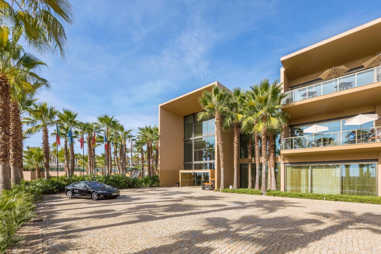 Ap Lago Montargil - Conference & Spa Exterior photo The photo shows a modern building with a sleek design, featuring large glass windows and balconies. The structure is surrounded by a well-maintained outdoor area with palm trees, adding a tropical feel. In front of the building, there is a paved park